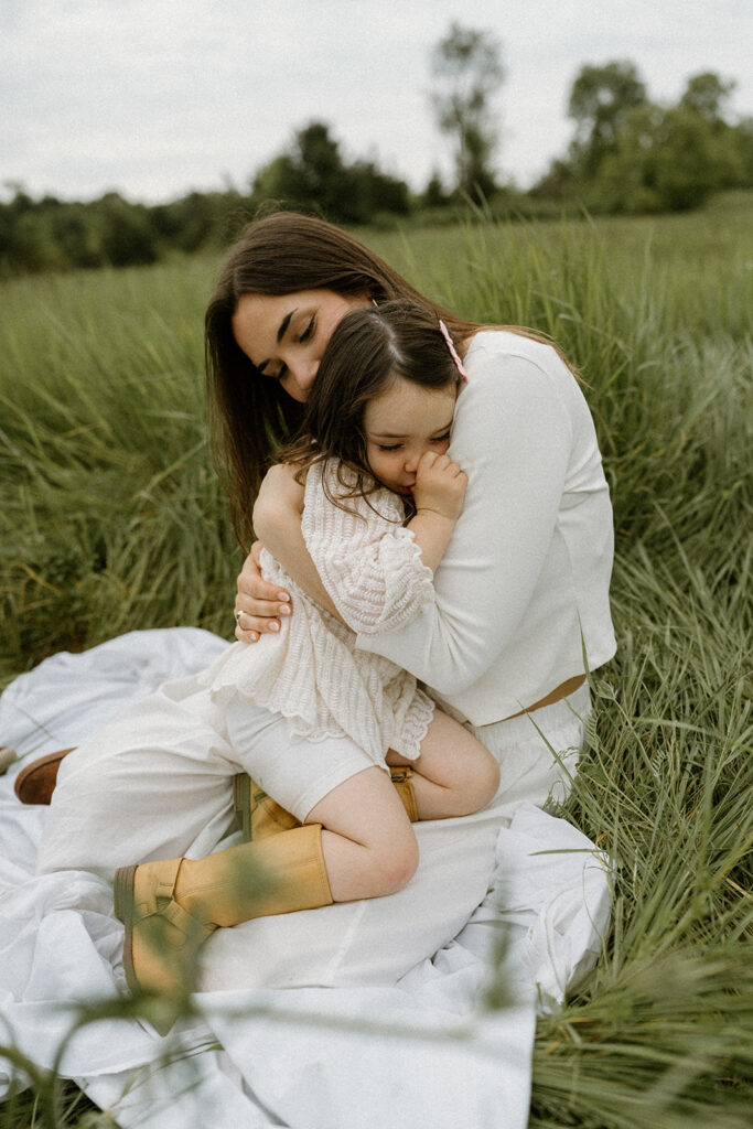 Mother hugging her young daughter while sitting on a blanket in tall grass during an outdoor Vermont session, a tender moment showing what to wear for family photos with soft neutral textures and natural tones.
