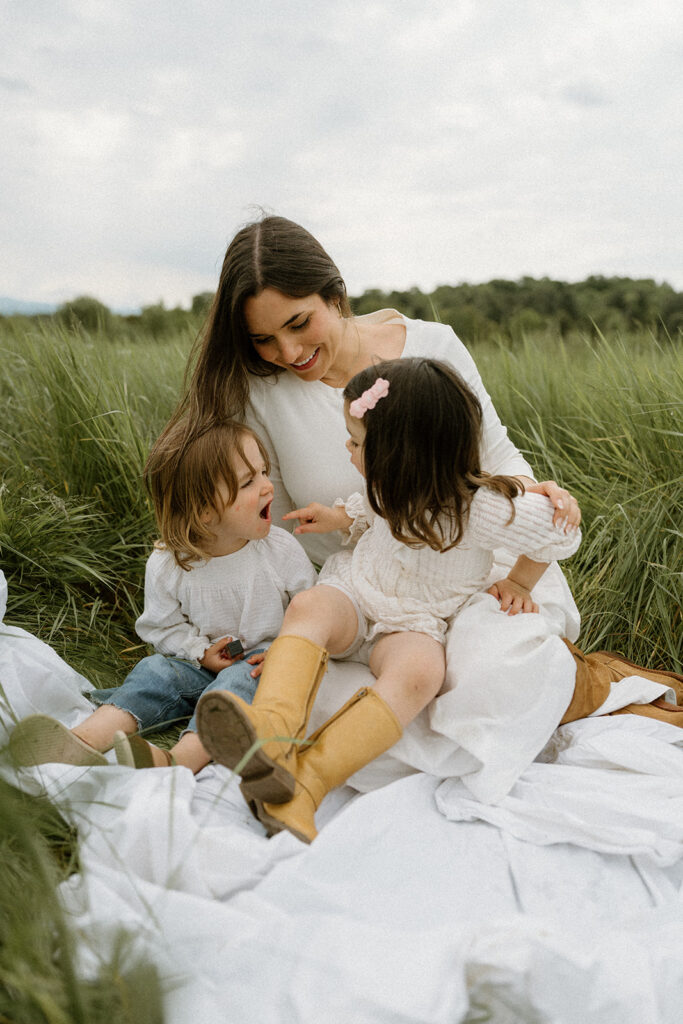 Mother sitting with her two young children on a blanket in a grassy field, photographed by a Vermont family photographer with relaxed neutral family photo outfits that complement the landscape.