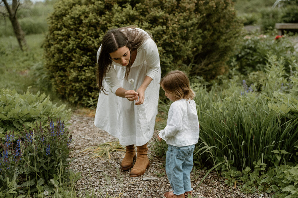 Mother showing her toddler a dandelion while walking through a garden path, a candid moment illustrating what to wear for family photos with soft neutral clothing and natural textures.