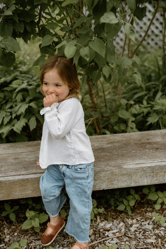 Young child standing beside a garden bench in simple denim and a soft white top, a natural example of comfortable outfits when you dress for family photos with young kids.