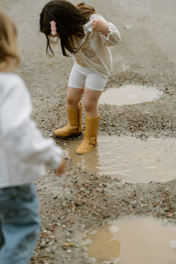 Child standing in a muddy puddle wearing yellow boots and a textured white dress, a playful moment often captured in relaxed family photo outfits.