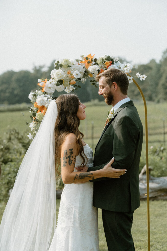 Bride and groom holding each other beneath a gold ceremony arch decorated with white and orange flowers overlooking a quiet Vermont countryside field.