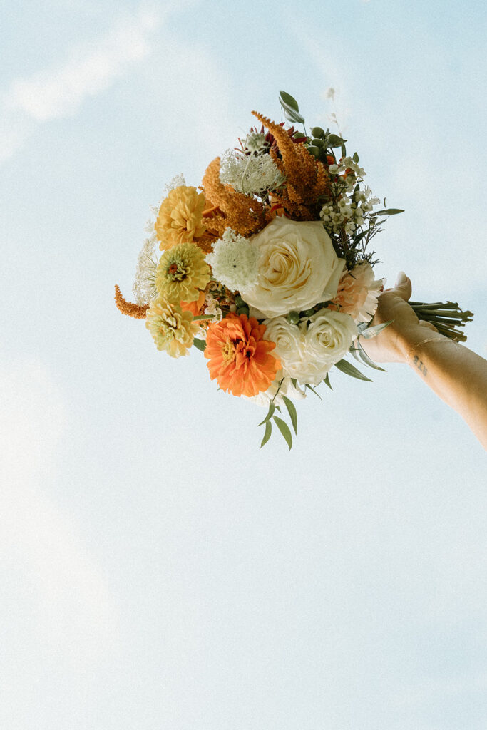 Colorful summer bridal bouquet with white roses, orange zinnias, and soft greenery held up against a pale sky.