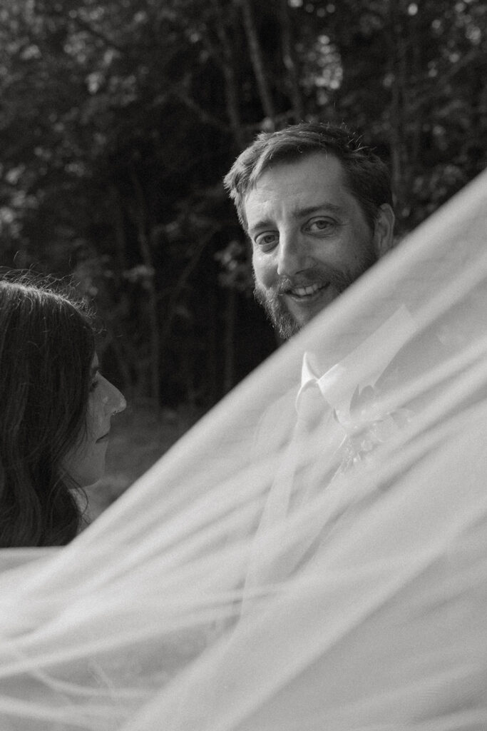 Black and white portrait of the groom smiling at the bride as her veil drifts across the frame during quiet wedding portraits.