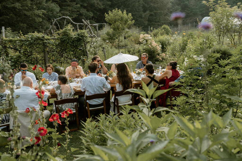 Guests gathered around a long wooden table for dinner surrounded by wildflowers and garden beds at an intimate backyard wedding in Vermont.