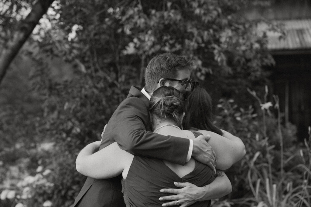 Black and white photo of the groom hugging two loved ones during an emotional moment at the outdoor wedding reception.