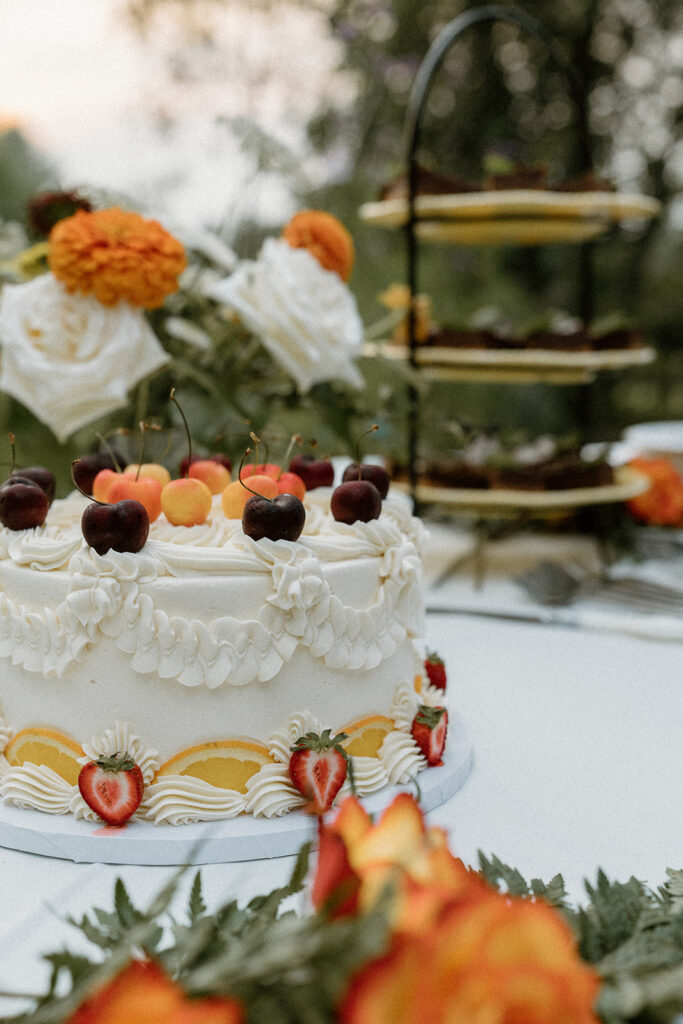 Vintage style wedding cake topped with cherries, strawberries, and orange slices displayed on an outdoor dessert table with flowers.