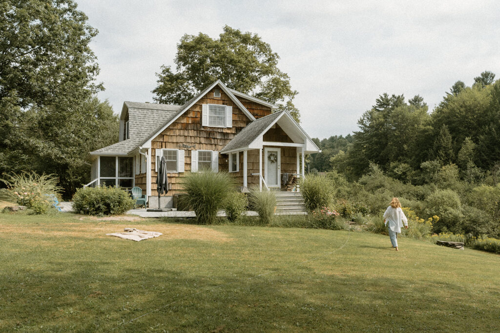 Small Vermont home surrounded by greenery and open yard where a mother walks across the lawn during lifestyle family photos at home.