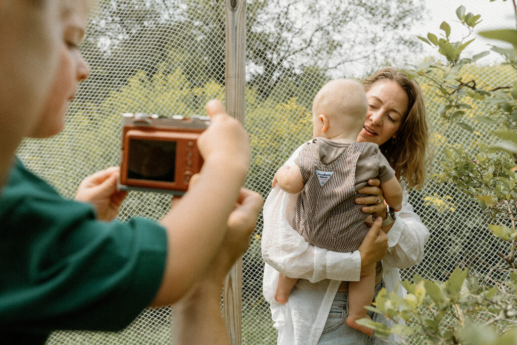 Young child photographing his mom and baby sibling with a tiny camera during playful lifestyle family photos in a backyard garden.