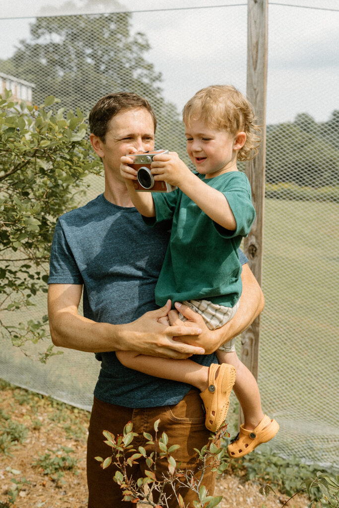 Dad holding his son while the toddler takes photos with a small camera during candid lifestyle family photos at home.