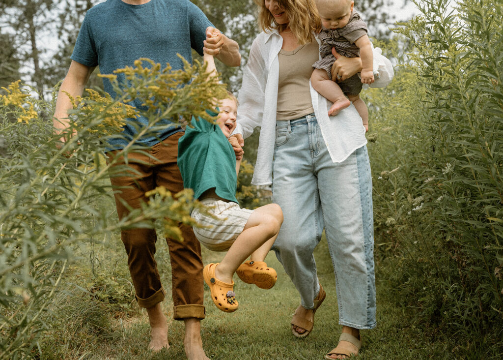 Parents walking through tall summer grass while swinging their laughing toddler during candid lifestyle family photos in Vermont.