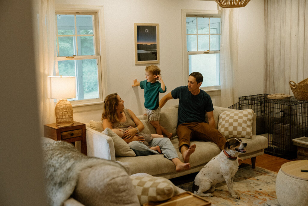 Candid lifestyle family photos of a Vermont family on their living room couch as their toddler stands between parents while the baby rests in mom’s arms.