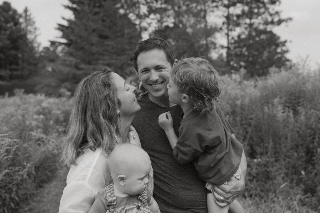 Black and white lifestyle family photo of parents smiling at each other while holding their children during a quiet outdoor moment.