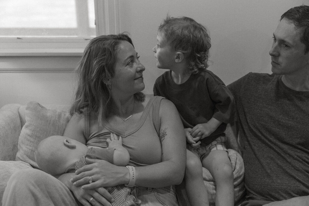 Mother holding baby while toddler leans in and father watches during quiet family moment on the couch.