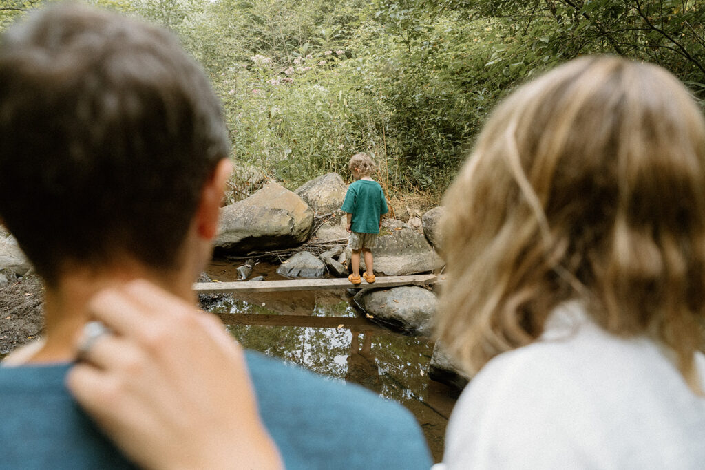 Young child balancing on rocks beside a small Vermont creek while parents watch nearby.