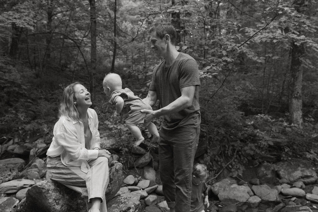 Black and white lifestyle family photo of parents playing with their baby beside a forest creek while their toddler explores nearby.