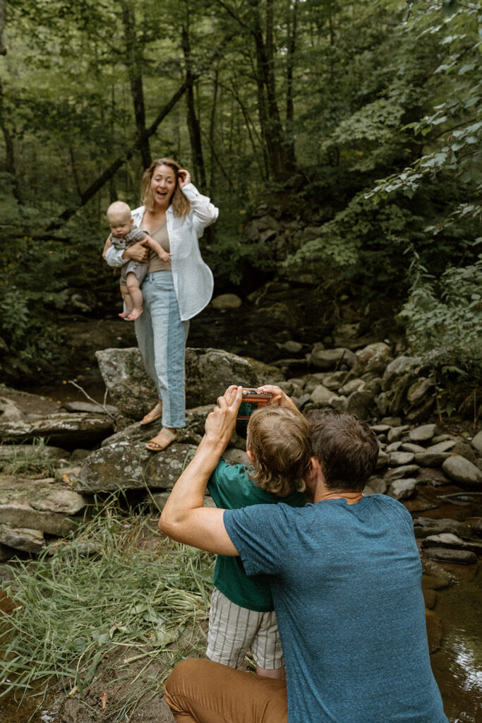 Dad helping his toddler take a photo of mom holding the baby by a wooded stream in Vermont