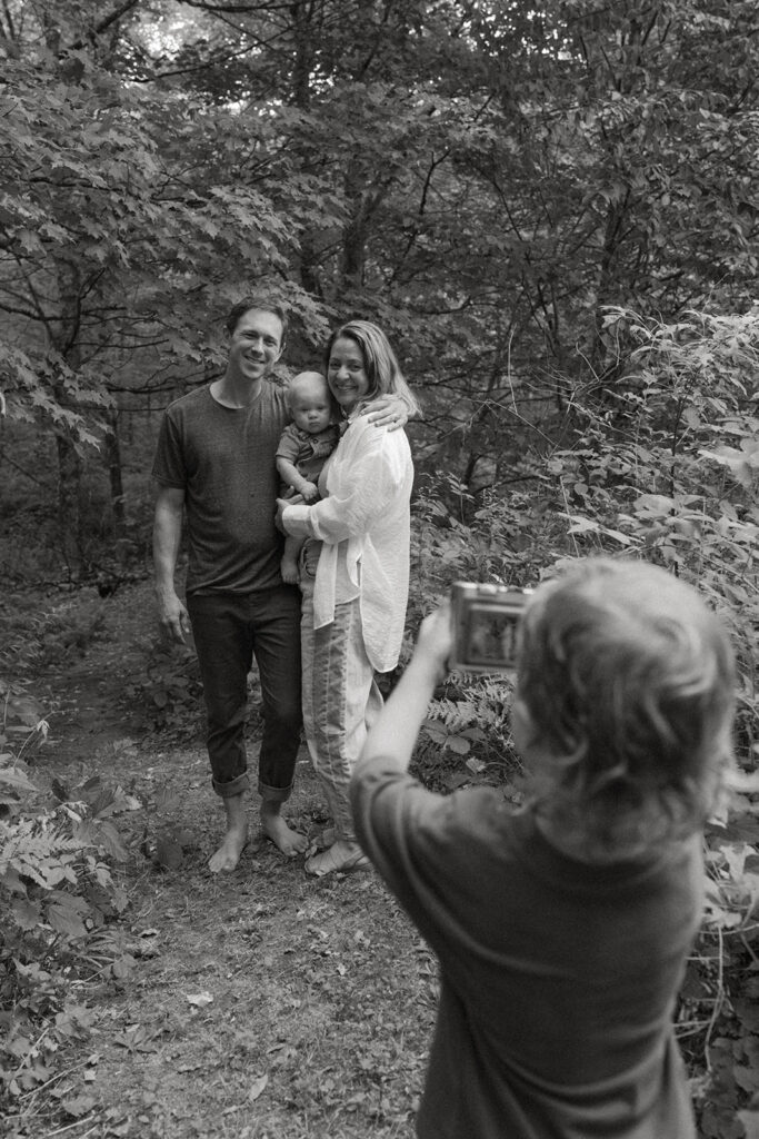 Black and white candid moment of toddler photographing parents holding baby during creative lifestyle family photos in the Vermont woods.