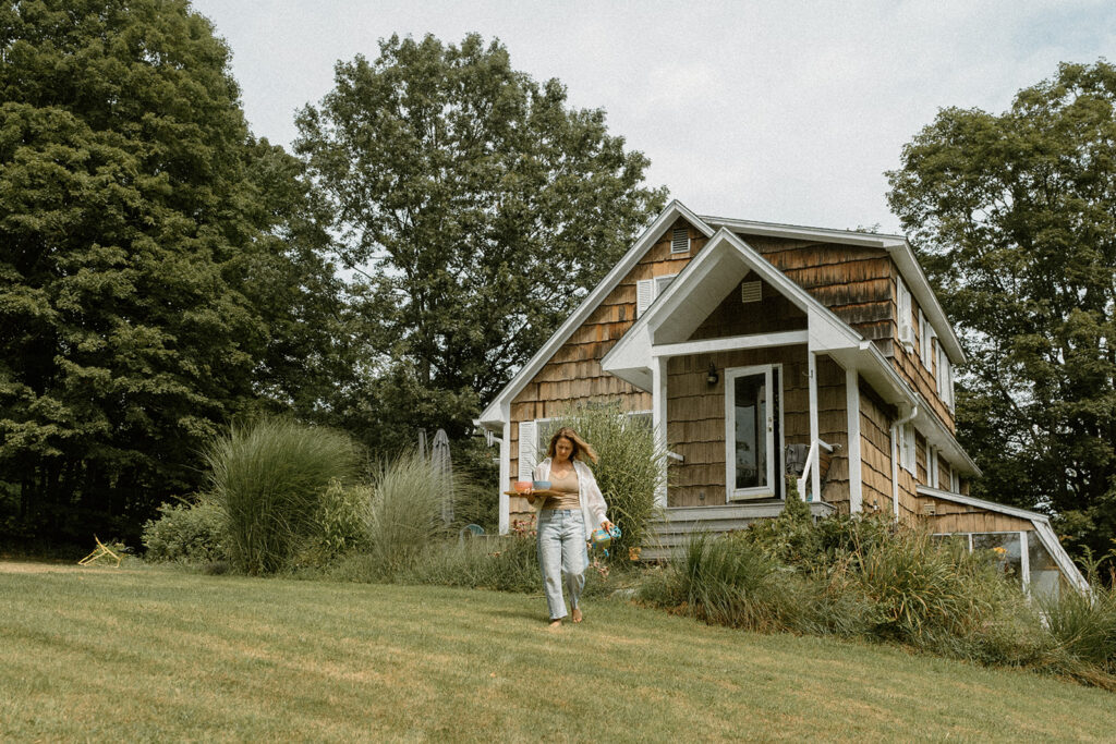 Small Vermont home surrounded by greenery where a mother walks down the yard with snacks in hand.
