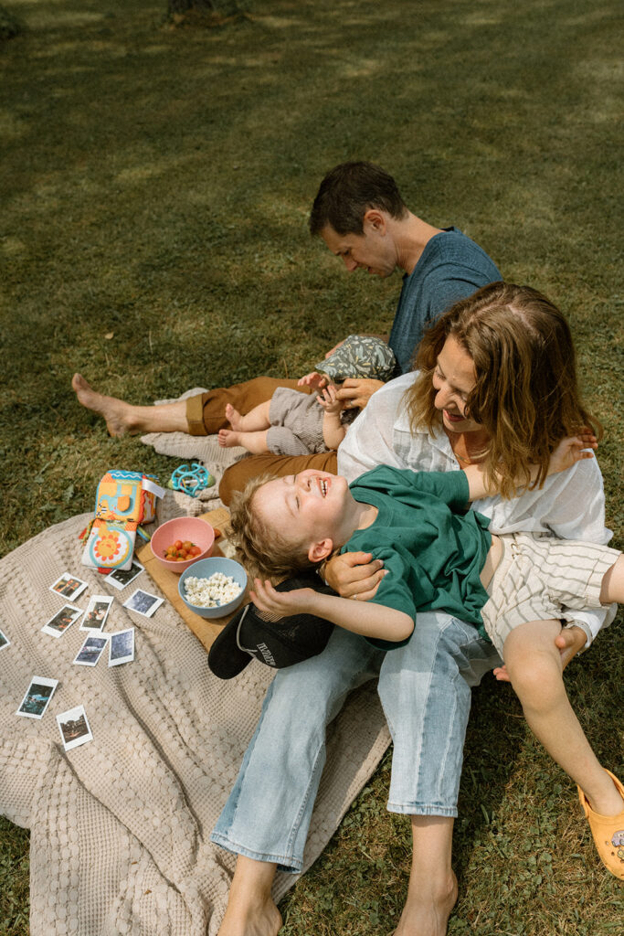 Mother laughing while her young son reclines across her lap during warm Vermont lifestyle family photos on a backyard picnic blanket.