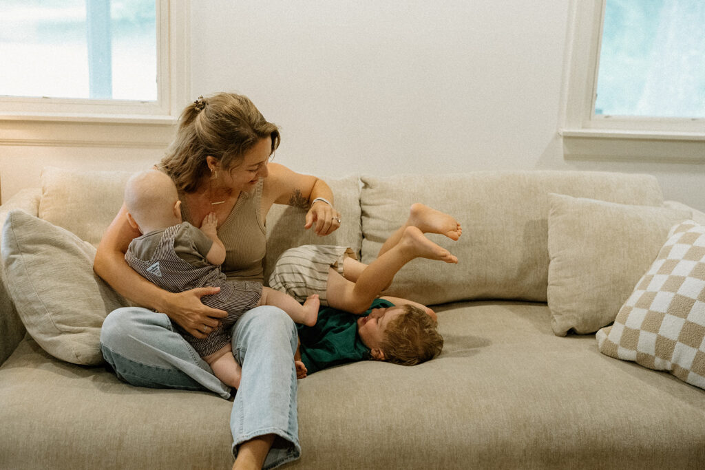 Mother sitting on a couch holding her baby while her young son playfully tumbles beside her.