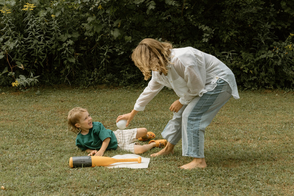 Candid lifestyle family photo of mom tossing a ball to her laughing toddler during playful backyard family photos.