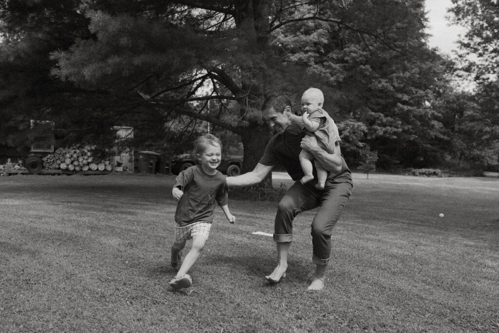 Black and white family photo of father holding baby while running with toddler across grassy Vermont yard.