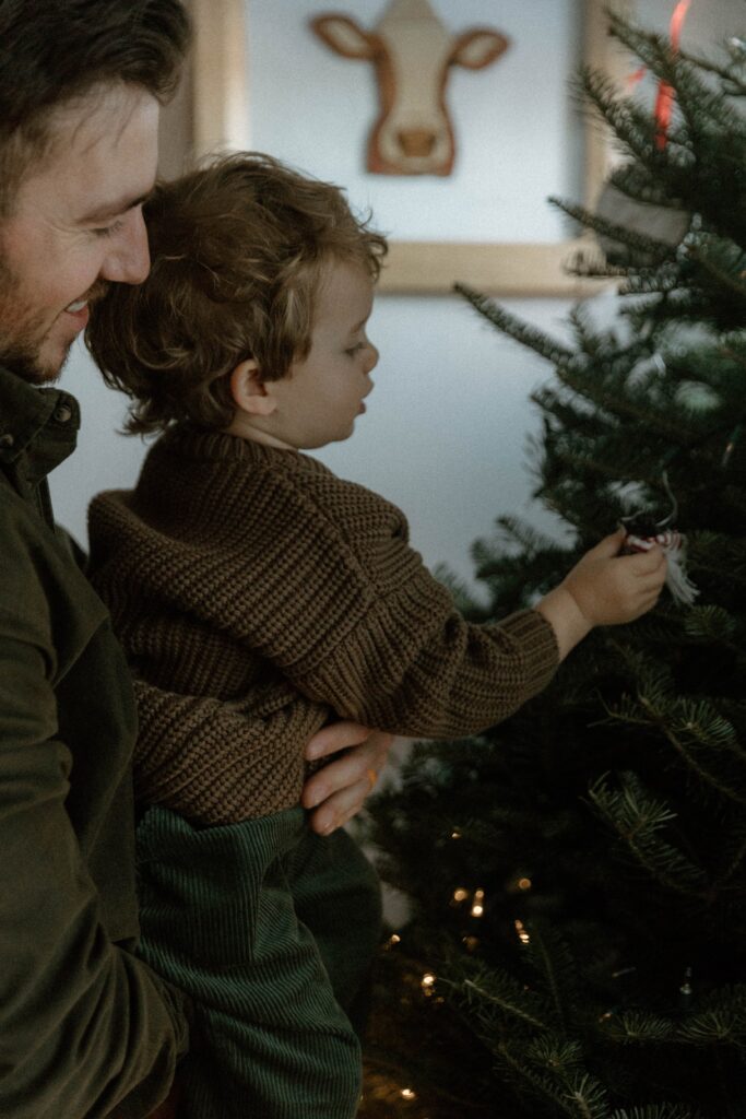 Father holding his toddler while decorating a Christmas tree, wearing relaxed and coordinated clothing that works well when you dress for family photos with comfort and movement in mind.