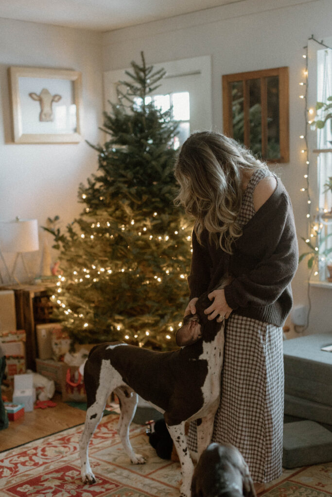 Mother in a neutral dress and cardigan interacting with her dog beside a Christmas tree, illustrating a warm family photos color scheme with layered textures and natural tones.