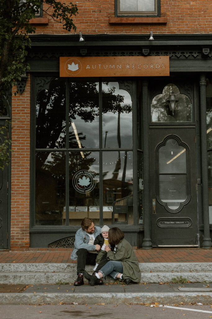 Family sitting on the sidewalk outside Autumn Records in Winooski, Vermont during a lifestyle session photographed by a Vermont family photographer.