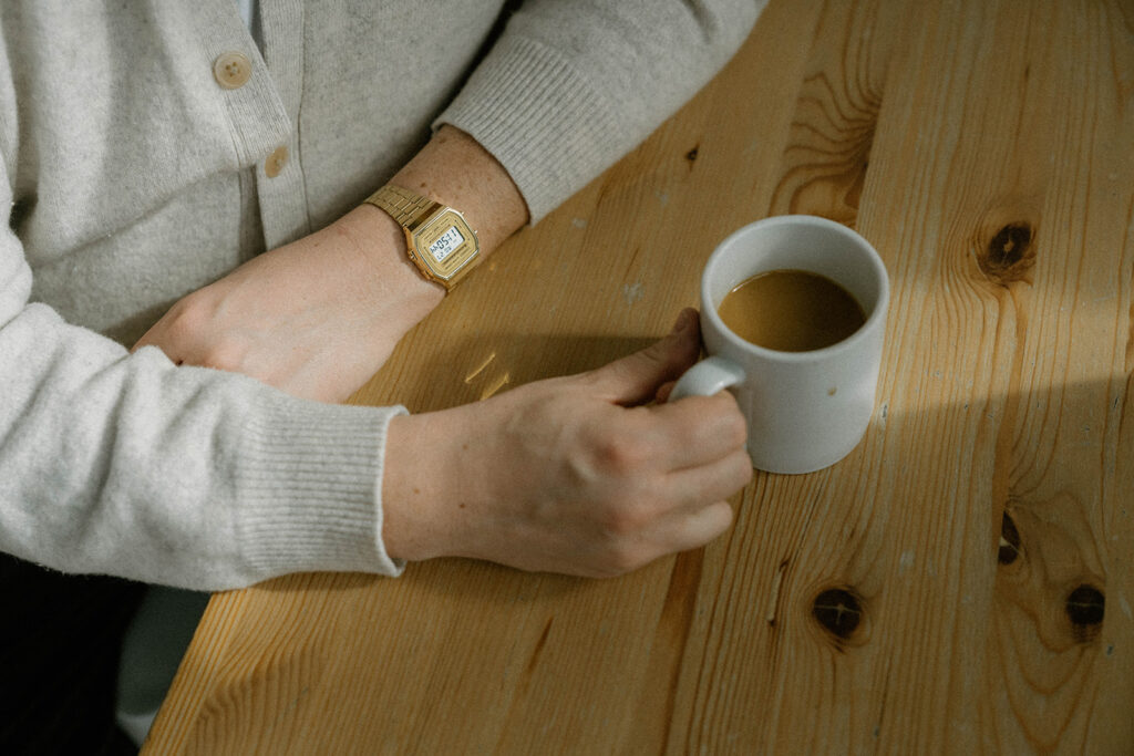 Close-up of a parent holding coffee at a wooden table in soft neutral clothing, a quiet lifestyle moment that reflects a relaxed family photos color scheme.