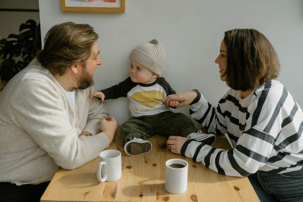 Parents sitting with their baby at a wooden table during an in-home lifestyle session, photographed by a Vermont family photographer with simple coordinated outfits.