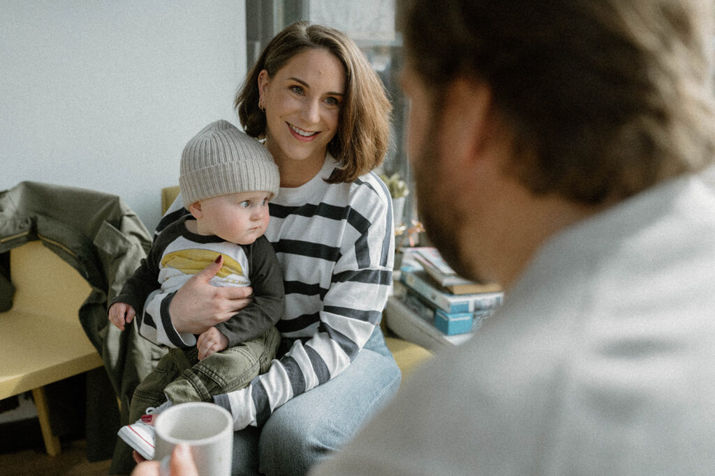 Mother holding her baby while smiling across the room during an in-home session, a natural example of what to wear for family photos with soft textures and everyday clothing.