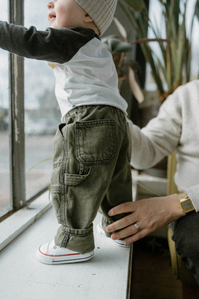 Baby standing at a window in green pants and white sneakers while a parent steadies them, illustrating a comfortable family photos color scheme with soft neutrals and earthy tones.