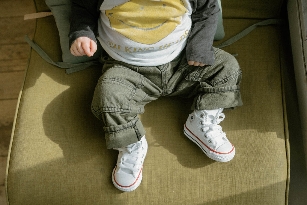 Baby wearing tiny Converse sneakers and green pants sitting on a chair, a playful detail of comfortable family photo outfits that allow kids to move naturally during a session.