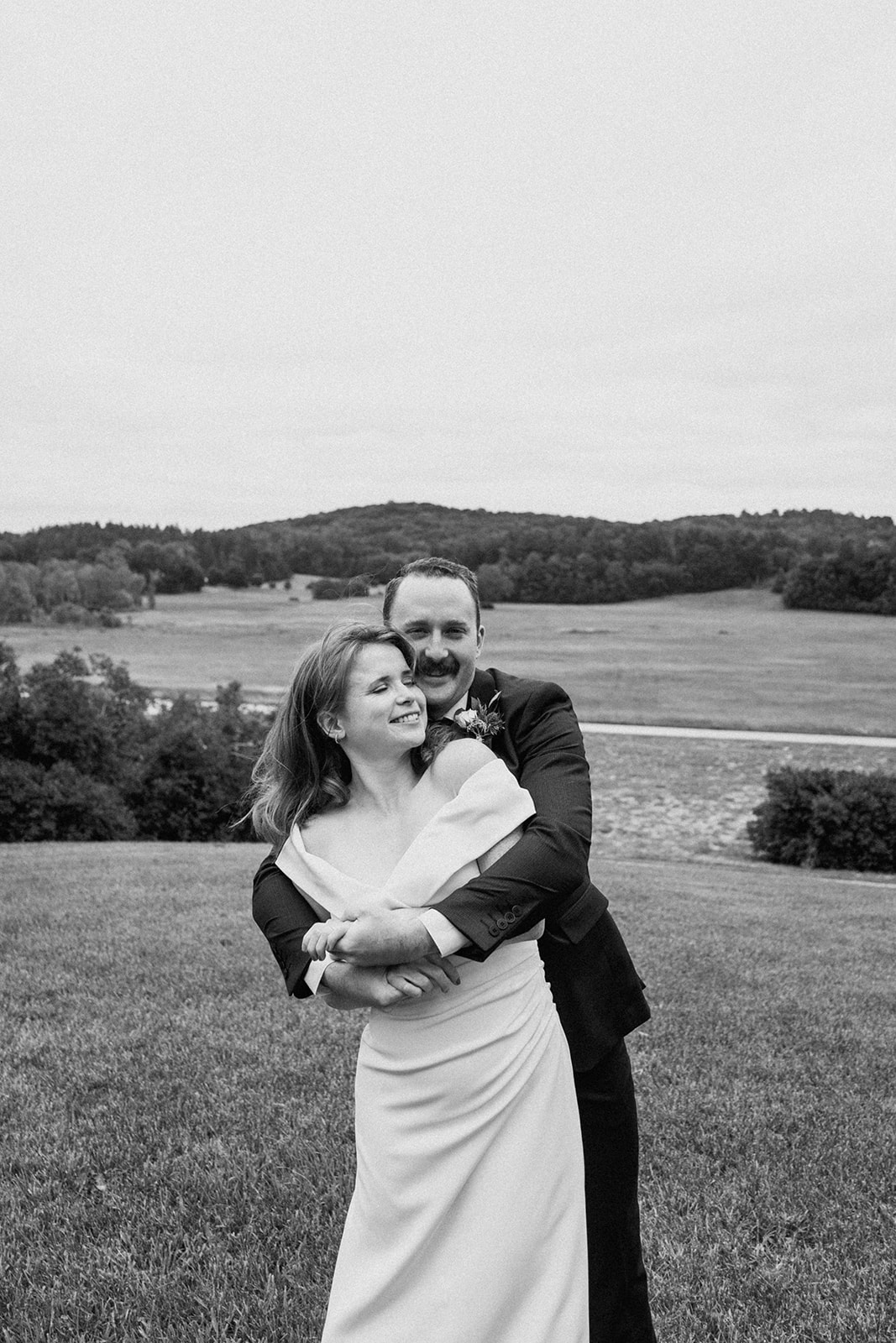 groom standing with arms around bride overlooking scenic view of Shelburne Farms in Vermont