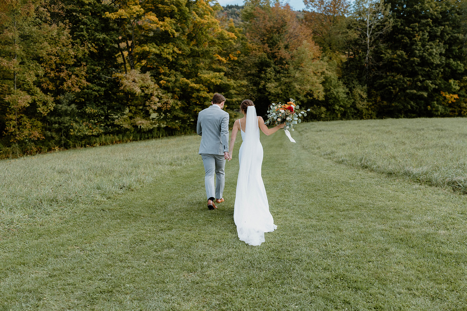 bride and groom walking through tree-lined field after fall elopement ceremony in Vermont