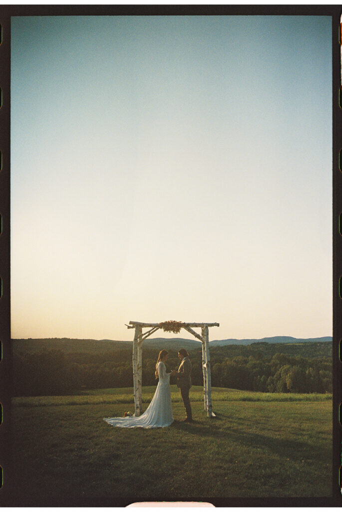 Bride and groom exchanging vows beneath a wooden arbor overlooking Vermont mountains at sunset, photographed by an intimate wedding photographer at a scenic Vermont wedding venue.