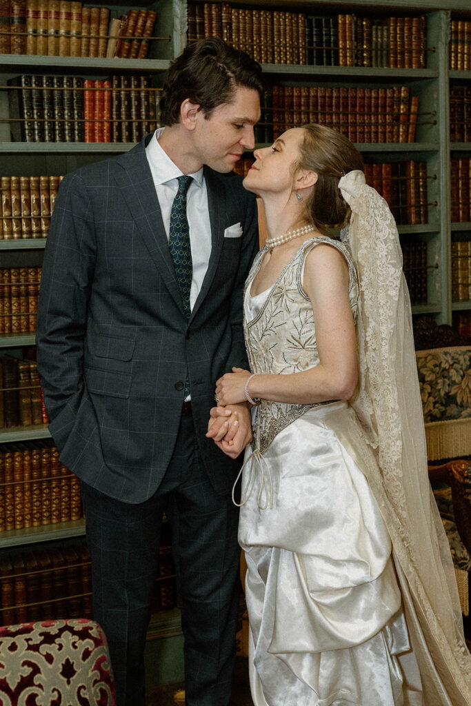 Bride in heirloom silk wedding dress and lace veil holding hands with groom in historic library at the Brick House, Shelburne Museum wedding in Vermont.