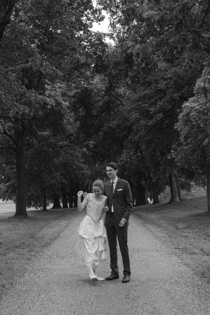 Bride lifting the hem of her heirloom wedding dress while walking with groom down the tree-lined drive at Shelburne Museum during an intimate Vermont wedding.