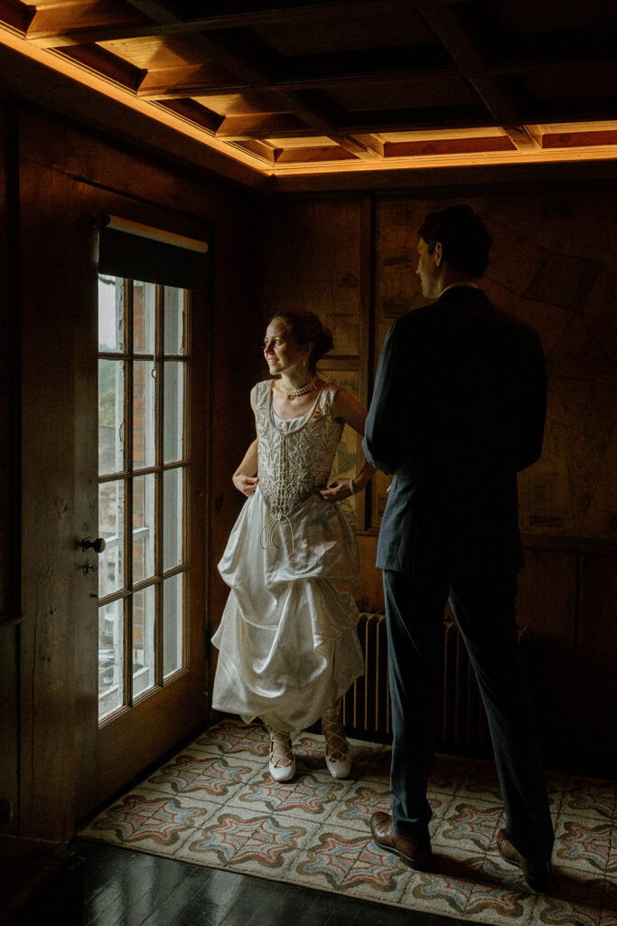 Bride in heirloom silk wedding dress and pearl necklace standing by window light with her groom inside the historic Brick House at Shelburne Museum.