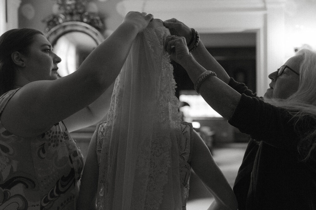 Mother and family member helping bride place heirloom lace veil before ceremony at Brick House wedding in Shelburne, Vermont.
