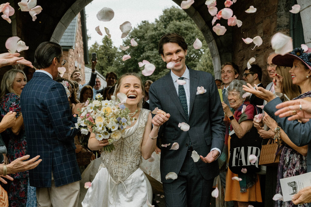 Bride and groom laughing while guests toss rose petals outside the Brick House ceremony at Shelburne Museum wedding in Vermont.