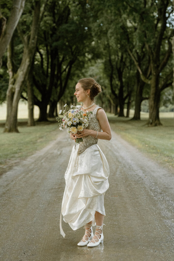 Bride holding bouquet while standing on the tree-lined drive at the Brick House, Shelburne Museum — an iconic intimate Vermont wedding location.