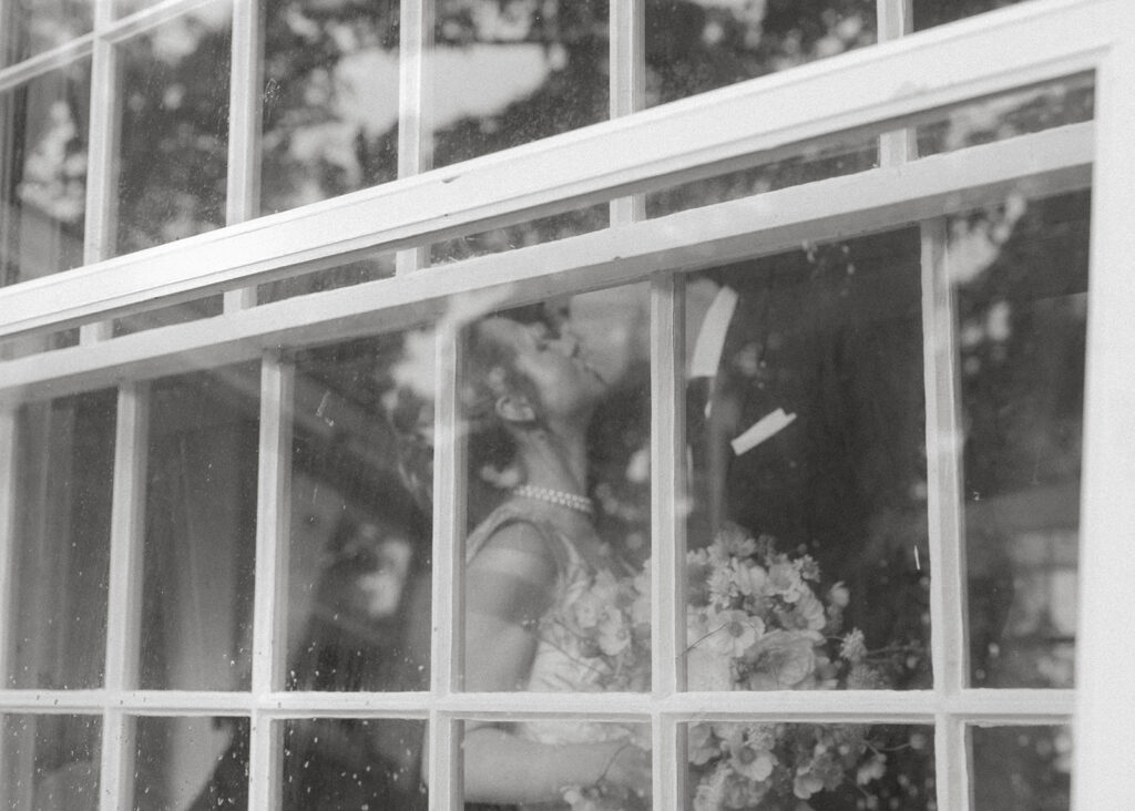 Bride holding bouquet and kissing groom reflected through historic window panes at the Brick House during romantic Vermont museum wedding.
