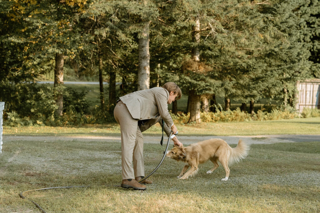 Groom playfully spraying water at his dog outside a farmhouse during a relaxed Vermont elopement day at one of the state’s intimate wedding locations.