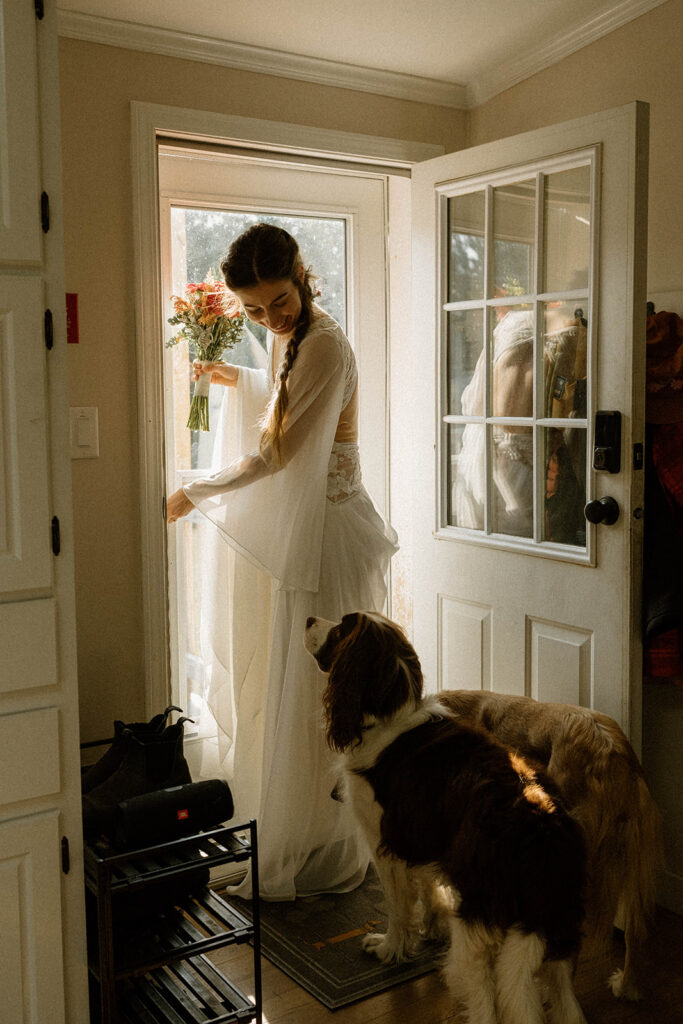 Bride holding her bouquet by a sunlit doorway while her dog waits beside her, a quiet getting-ready moment