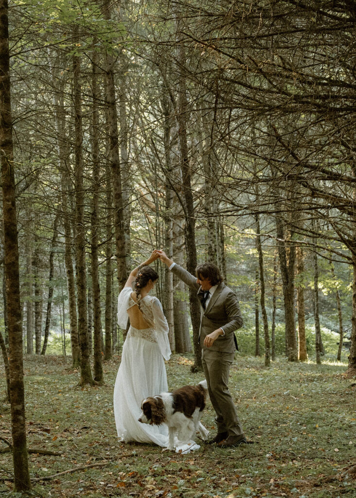 Newly married couple twirling together in a pine forest as their dog runs between them during their Vermont elopement at one of the region’s most beautiful intimate wedding locations.
