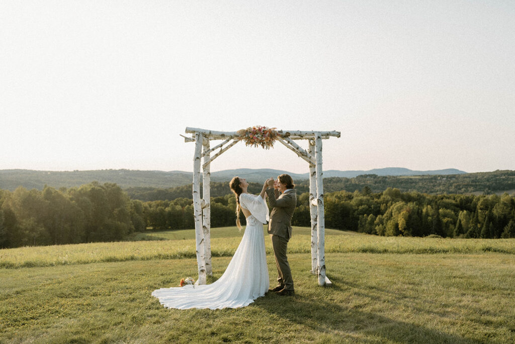 Bride and groom laughing beneath a wooden arbor overlooking Vermont mountains during their intimate wedding ceremony.