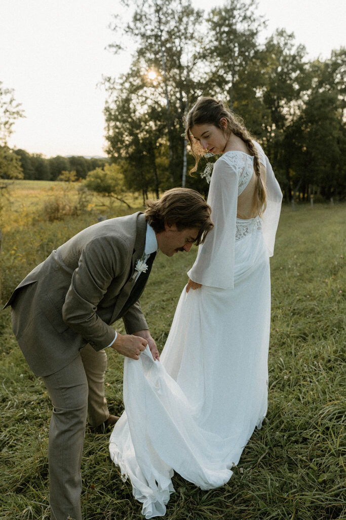 Groom adjusting the bride’s dress in a grassy field during a Vermont intimate wedding photographed by a documentary-style elopement photographer.